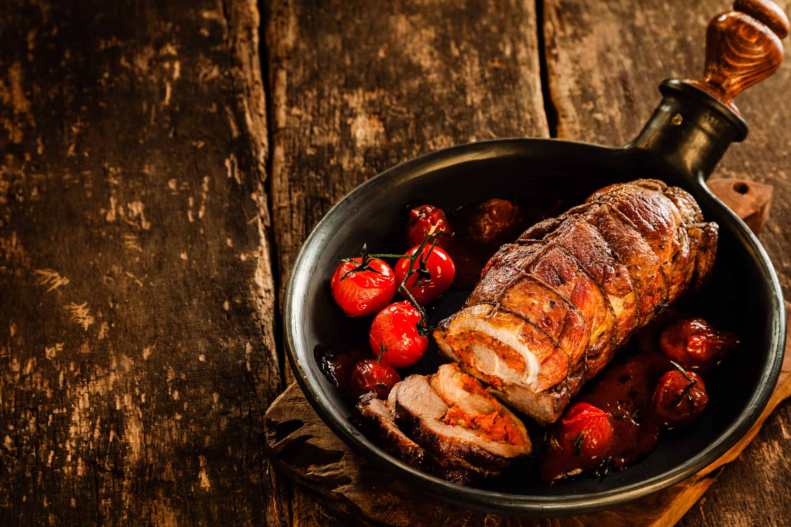 High Angle View of Gourmet Roast Pork Roll Stuffed with Tomatoes and Grilled Vegetables and Served on Sizzling Cast Iron Pan on Rustic Wooden Table with Copy Space